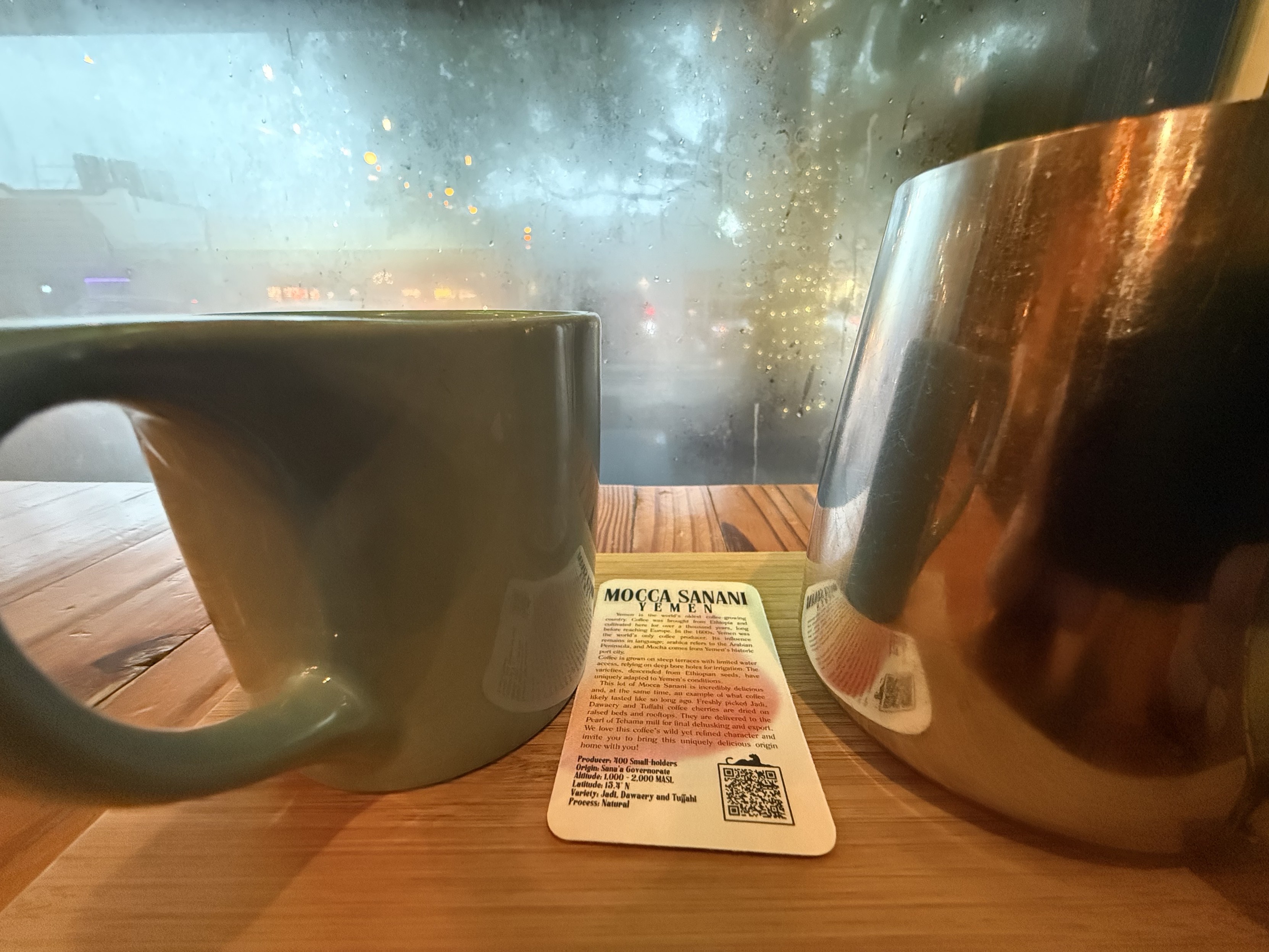 A tasting card for panther’s Yemeni “Mocca Sanani” coffee between a green ceramic mug and a small metal pitcher, with a condensation-covered window and rainy morning traffic in the background