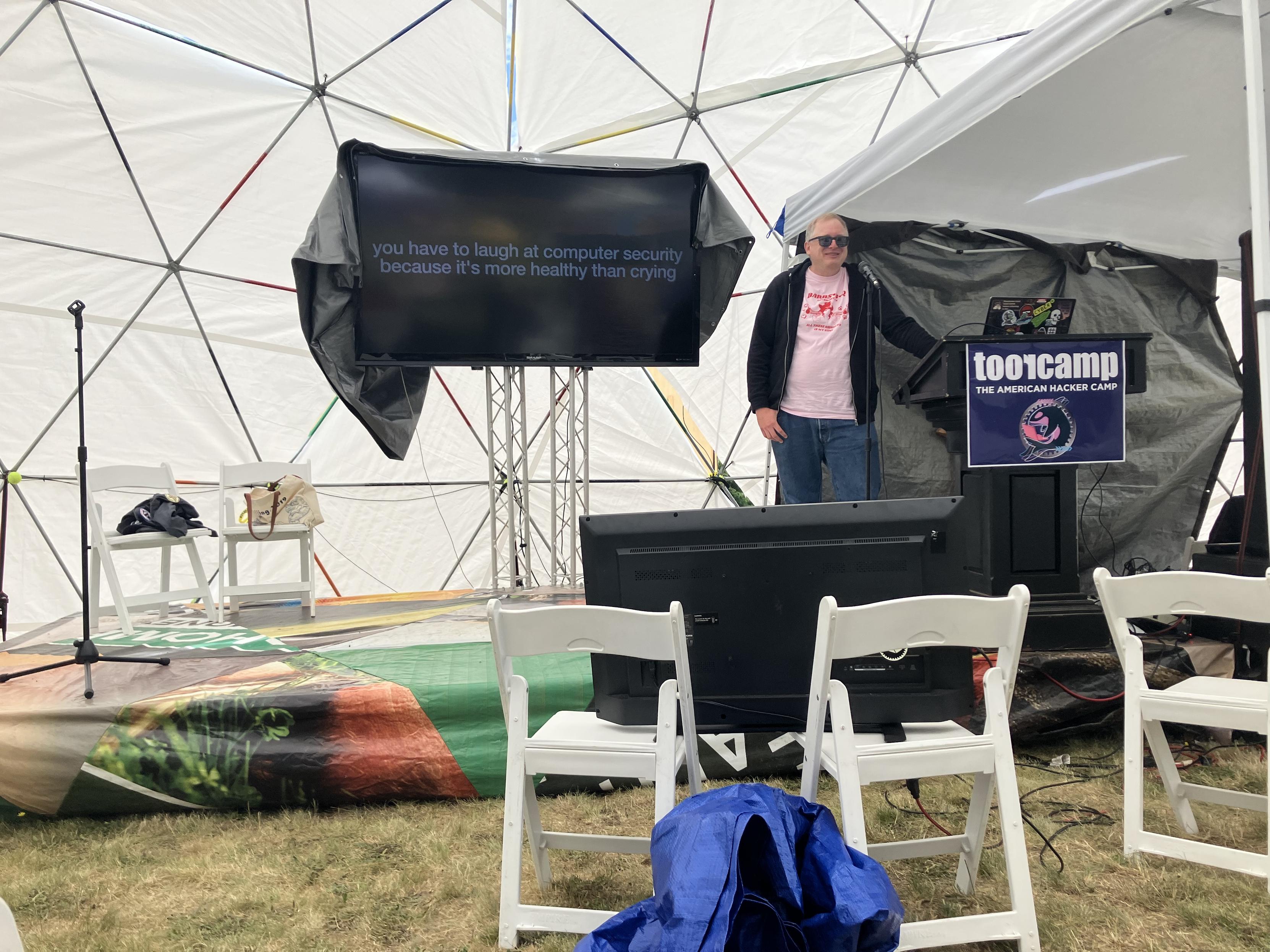 I’m in stage in a white dome, next to a screen that says “you have to laugh at computer security because it’s more healthy than crying”